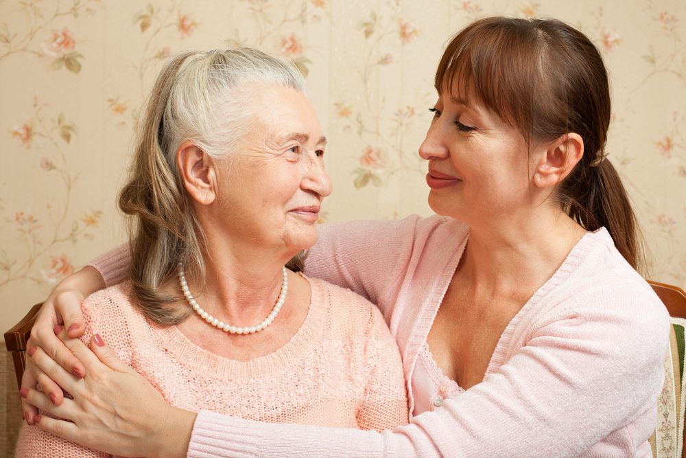 mamma anziana con sua figlia di mezza età, sorridente e abbracciata, con indosso camicette rosa chiaro.