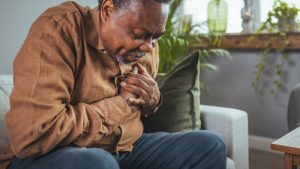 Uomo seduto sul divano di casa, mano sul petto mentre avverte un dolore toracico che potrebbe essere la sindrome del cuore spezzato