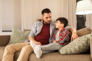 Padre e figlio seduti sul divano marrone chiaro a casa, parlando insieme